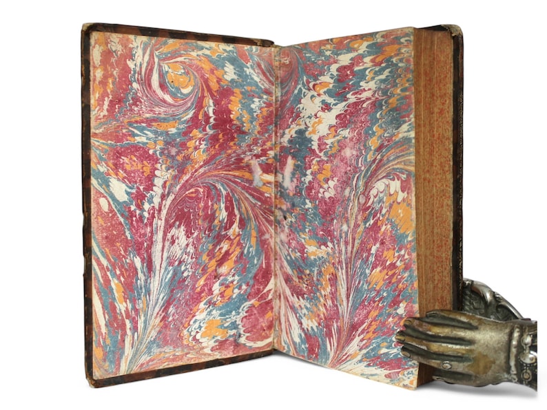 May include: An open antique book with marbled pages in shades of red, blue, yellow, and white. The book's cover is dark brown, and the pages are held open by a decorative metal hand. The book's pages have a swirling pattern.