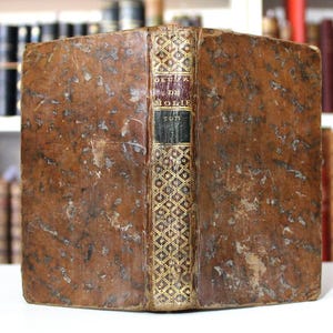 K&ouml;nnte beinhalten: Antikes Buch mit einem abgenutzten, braunen Ledereinband. Der Buchr&uuml;cken zeigt goldene Verzierungen und den Titel "OEUVRE DE MOLIERE TOME". Das Buch wird vor einem verschwommenen Hintergrund anderer B&uuml;cher pr&auml;sentiert.