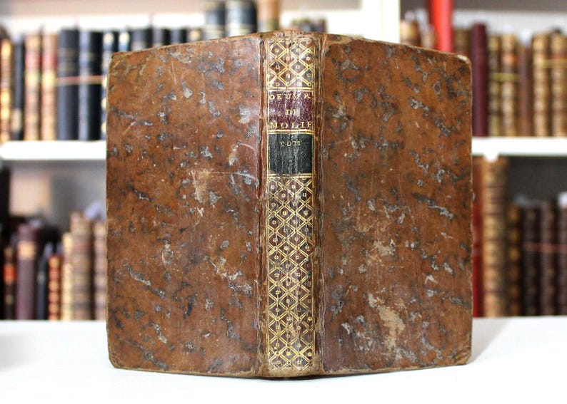 K&ouml;nnte beinhalten: Antikes Buch mit einem abgenutzten, braunen Ledereinband. Der Buchr&uuml;cken zeigt goldene Verzierungen und den Titel "OEUVRE DE MOLIERE TOME". Das Buch wird vor einem verschwommenen Hintergrund anderer B&uuml;cher pr&auml;sentiert.