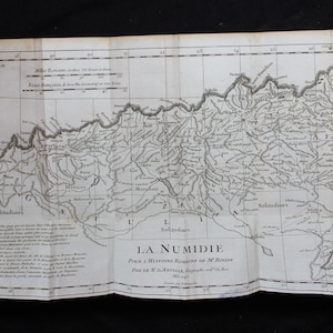 May include: Antique map of La Numidie, featuring detailed geographical features and text in French. The map is folded, showing creases and age-related wear. The map's title, "LA NUMIDIE," is prominently displayed, indicating its subject matter.