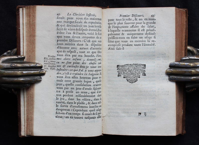 Pode incluir: Um livro aberto com um desenho decorativo na p&aacute;gina direita. O texto na p&aacute;gina esquerda diz: "Le Chrisien inftrvis, feroit pour vous des maintenan une marque fatale de reprobatin, & qui deviendroit un jour le t fujet de votre defeſpoir &eacute;ternel. Por &eacute;viter l'un & l'autre, voici le fut que vous devez empotter de a premier Difcours: C'est que vou vous mettiez dans la difpofisis d'&eacute;couter avec autant d'attentiu que de refpect, tout ce que Din Venite vous dira par ma bouche. Fo Fin, audite mes chers enfans &eacute;coutez mi mco ne font point des choses so ni docebo nes & curieuses dont je veux vo Pial inftruire ce que j'ai &agrave; vous appro dre, c'est &agrave; craindre le Seigneur. vous &ecirc;tes allez heureux pour tenir cette grande le&ccedil;on, quri joye, quelle confolation n'art vous pas un jour d'avoir f&ccedil;um tre &agrave; profit un rems, que di tres perdent miferablement dati le jeu, dans les vifites, dans vanit&eacute;, dans le plaifir, && dans mi le fortes d'amufemens inutiles of dangereux Cependant quel ulaga fait-on d'un temps fi court & fi po cicux on en trouve to&ucirc;jours a"