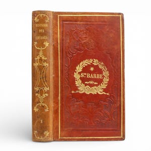 May include: An antique book with a red leather cover and gold detailing. The spine is tan with gold lettering and decorative patterns. The front cover displays a gold wreath design with the text "ST. BARBE".