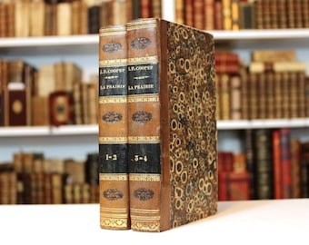 May include: Two antique books with brown leather covers and gold detailing. The spines are black with gold lettering and decorative patterns. The book on the right has a patterned cover. The books are labelled "L.P. Cooper" and "La Prairie."