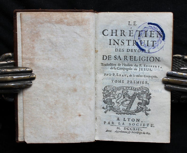 Pode incluir: Um livro antigo intitulado "Le Chretien Instruit des Devoirs de Sa Religion." O livro est&aacute; aberto na p&aacute;gina do t&iacute;tulo, que apresenta um bras&atilde;o decorativo e o texto "A Lyon, Par La Societe. M. DCCXII."