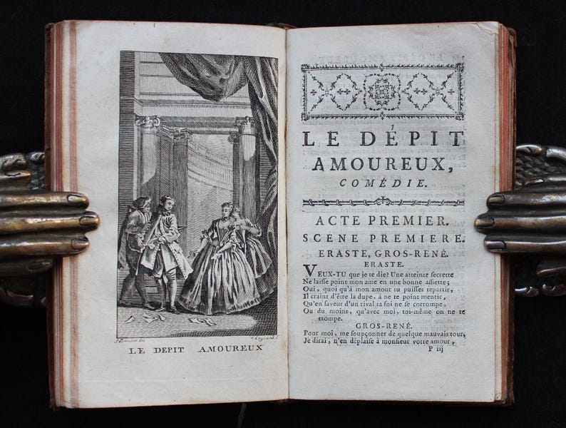 K&ouml;nnte beinhalten: Ein aufgeschlagenes antikes Buch mit einer Schwarz-Wei&szlig;-Illustration auf der linken Seite und franz&ouml;sischem Text auf der rechten Seite. Die Illustration zeigt drei Figuren in einem klassischen Rahmen. Der Buchtitel lautet "LE D&Eacute;PIT AMOUREUX."