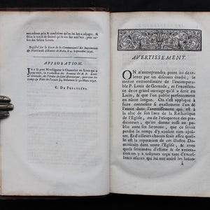 May include: An open antique book with aged, yellowed pages. The left page features text in French, including the word "APPROBATION." The right page also has French text, with the heading "AVERTISSEMENT." The book's binding is dark brown.