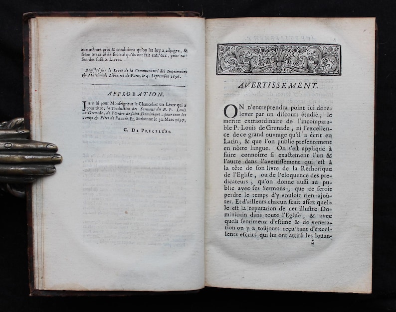 May include: An open antique book with aged, yellowed pages. The left page features text in French, including the word "APPROBATION." The right page also has French text, with the heading "AVERTISSEMENT." The book's binding is dark brown.