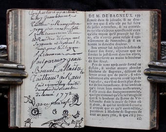 以下が含まれることがあります： 2ページ開かれたアンティークの本。左ページには1772年と記された黒インクの手書きの筆記体があり、下部には太陽、月、動物の小さな手描きのイラストがある。右ページにはフランス語の印刷されたテキストが記載されている。