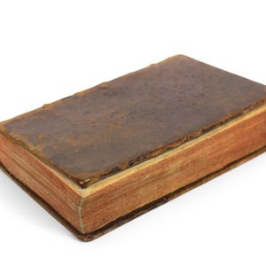 Pode incluir: Um livro vintage encadernado em couro marrom com p&aacute;ginas envelhecidas. O livro est&aacute; fechado e sobre uma superf&iacute;cie branca.