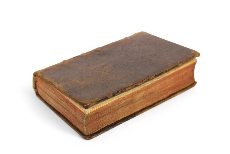 Pode incluir: Um livro vintage encadernado em couro marrom com p&aacute;ginas envelhecidas. O livro est&aacute; fechado e sobre uma superf&iacute;cie branca.