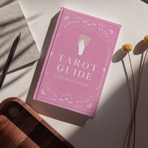 May include: A pink tarot guide book with the title "Tarot Guide f&uuml;r Anf&auml;nger" in white lettering. The book has decorative borders and a hand holding cards illustration. A pencil, notebook, and wooden tray are also visible.