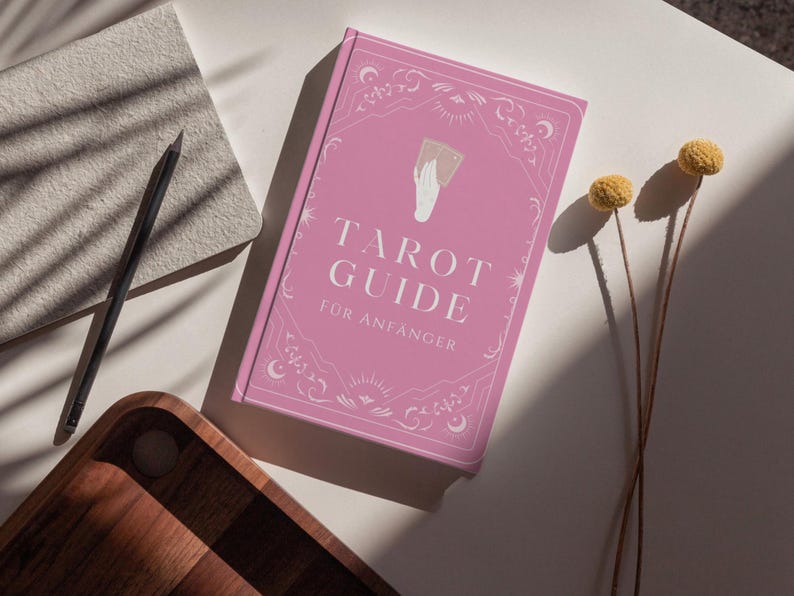May include: A pink tarot guide book with the title "Tarot Guide f&uuml;r Anf&auml;nger" in white lettering. The book has decorative borders and a hand holding cards illustration. A pencil, notebook, and wooden tray are also visible.
