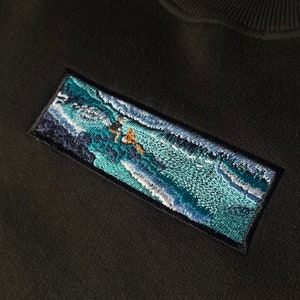 Op de afbeelding: Een zwart sweatshirt met een geborduurde patch die een blauwe en witte golf met een figuur die erop surft afbeeldt.