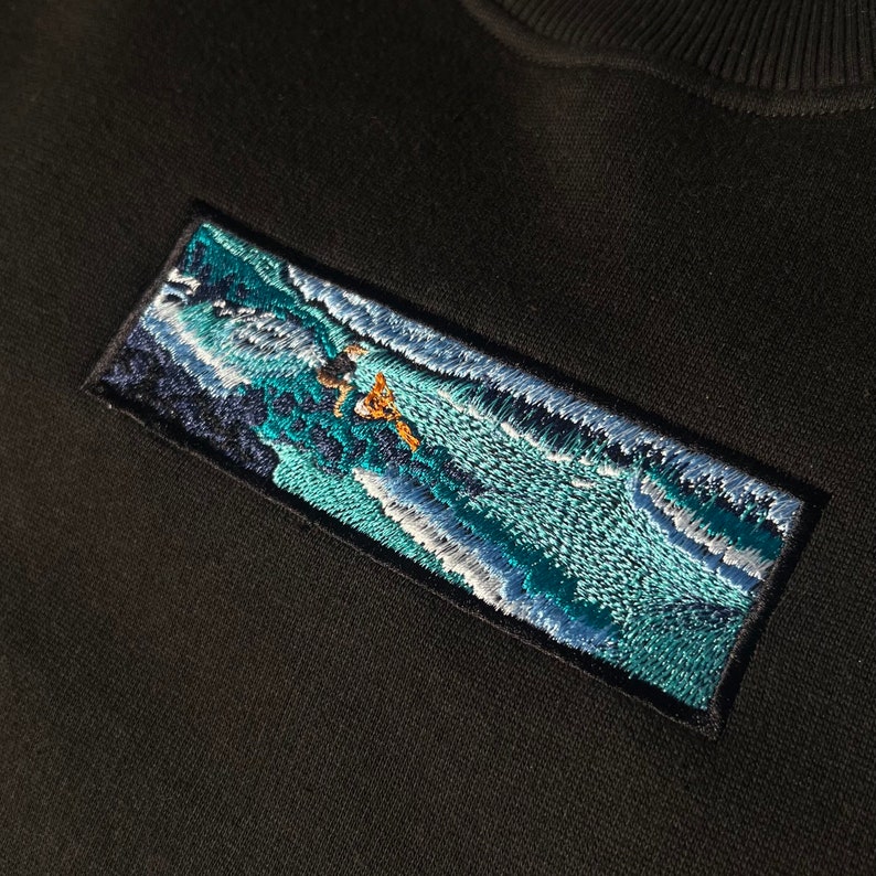 Op de afbeelding: Een zwart sweatshirt met een geborduurde patch die een blauwe en witte golf met een figuur die erop surft afbeeldt.