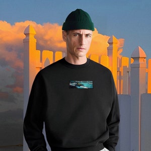 Op de afbeelding: Een zwart sweatshirt met een afbeelding van een zeelandschap op de voorkant. Het sweatshirt heeft een ronde hals en lange mouwen. Het model draagt een groene beanie.