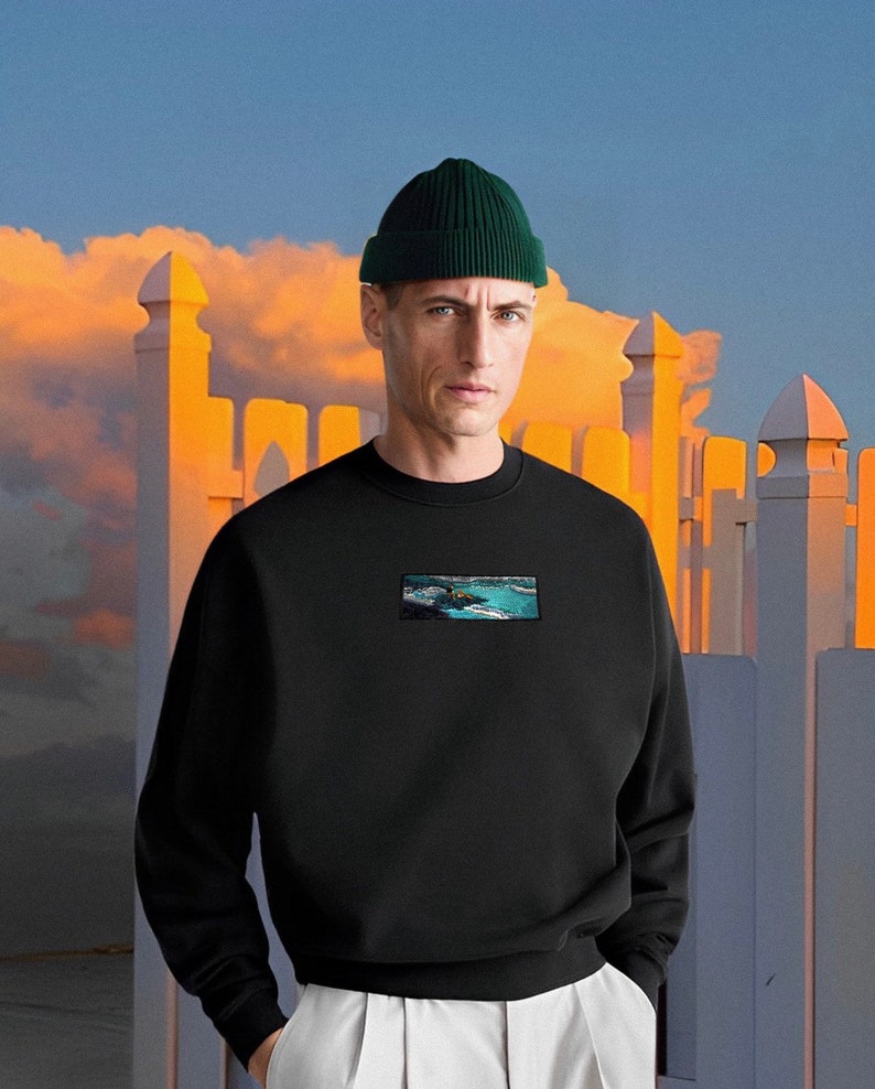 Op de afbeelding: Een zwart sweatshirt met een afbeelding van een zeelandschap op de voorkant. Het sweatshirt heeft een ronde hals en lange mouwen. Het model draagt een groene beanie.