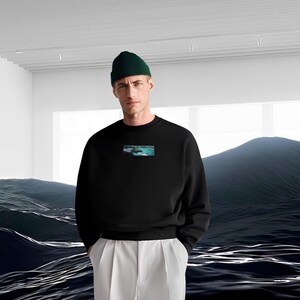 Op de afbeelding: Een zwart sweatshirt met een kleine grafische print van een zeelandschap op de voorkant. Het model draagt een groene beanie en een witte broek.