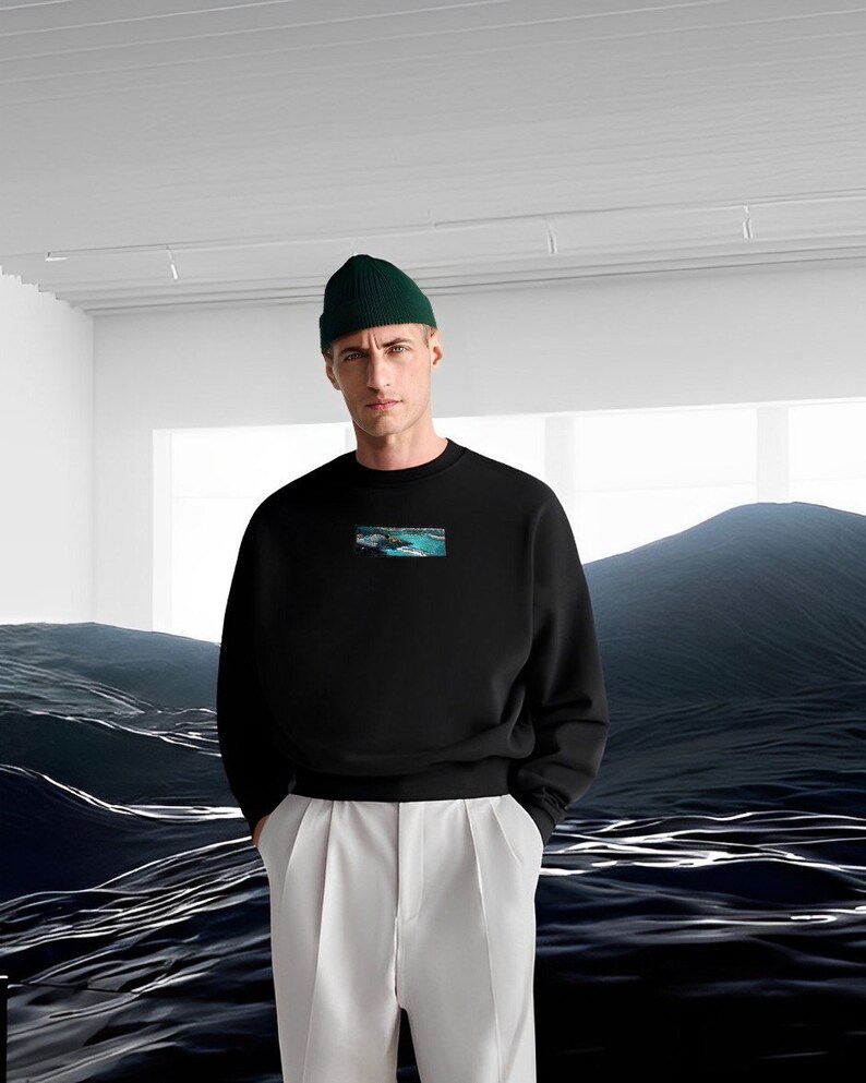 Op de afbeelding: Een zwart sweatshirt met een kleine grafische print van een zeelandschap op de voorkant. Het model draagt een groene beanie en een witte broek.