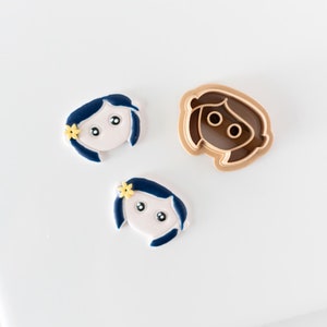 May include: Two blue and white fondant figures with yellow flowers and a brown plastic cookie cutter in the shape of a girl's head.