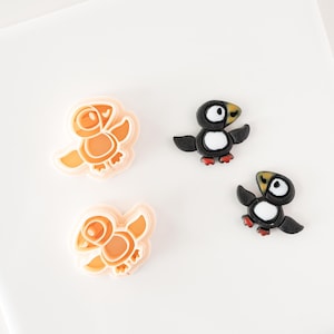 May include: Four plastic cookie cutters in the shape of puffins. Two are orange and two are black.