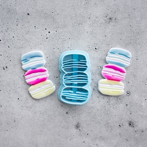 Puede incluir: Un juego de tres cortadores de galletas de macarons de color pastel y un molde de silicona para hacer macarons. Los cortadores están apilados uno encima del otro y el molde está en el centro.