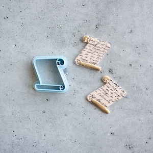 May include: A blue plastic cookie cutter shaped like a scroll, next to two fondant scrolls with gold accents and a handwritten script design.