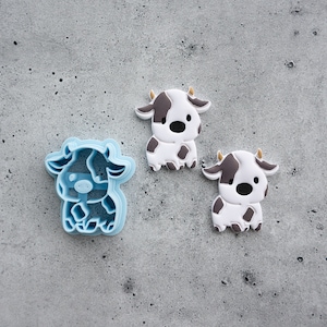 Op de afbeelding: Drie blauwe plastic koekjesvormen in de vorm van koeien. De koeien zijn wit met zwarte vlekken en hebben zwarte ogen en neuzen.