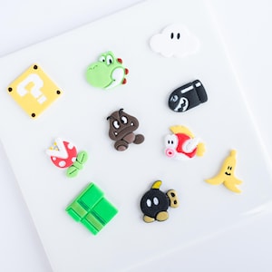 Puede incluir: Una colección de figuras de personajes de estilo caricatura coloridas, que incluyen un dinosaurio verde, una seta marrón, un signo de interrogación amarillo y un plátano amarillo. Las figuras están dispuestas sobre una superficie blanca.