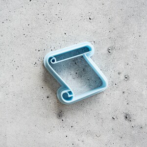 May include: A light blue plastic cookie cutter shaped like a scroll. The cutter is on a gray textured surface.