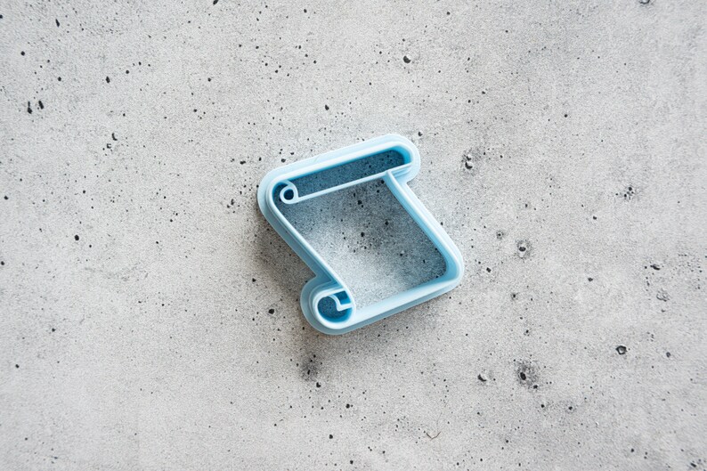 May include: A light blue plastic cookie cutter shaped like a scroll. The cutter is on a gray textured surface.