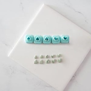 May include: A set of five light blue square-shaped clay cutters with distinct botanical-themed impressions on the faces, arranged in a horizontal row above ten small, light green clay shapes that mirror the designs of the cutters.
