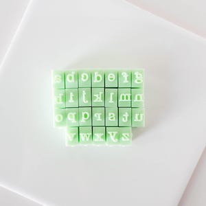 May include: A set of twenty-six light mint green rectangular rubber stamps, each featuring a single white lowercase letter of the alphabet from a to z, arranged in a grid formation with the letters oriented in reverse for stamping purposes.