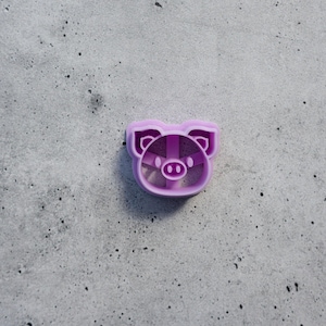 May include: A light purple plastic cookie cutter shaped like a pig face. The design features a rounded snout, two pointed ears, and internal structural bars that form the facial features, including the eyes and the outline of the pig's head.