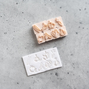May include: A set of eight small, light peach-colored rectangular stamps, each featuring a unique vegetable design, alongside a single white rectangular silicone mold that displays the corresponding vegetable impressions in a grid-like arrangement.