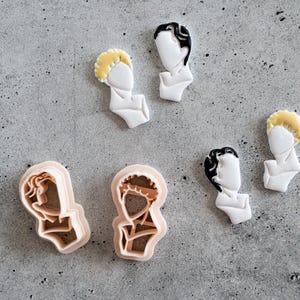 May include: A collection of face-shaped cookie cutters and cookies. The cookies have white faces with black or yellow hair. The cookie cutters are tan and white. The cookies and cutters are on a gray surface.