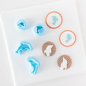 Könnte beinhalten: Ein Set mit blauen Delfin-förmigen Ausstechern und Fondant-Dekorationen. Die Fondant-Dekorationen befinden sich auf orangen Kreisen mit blauen Streifen.