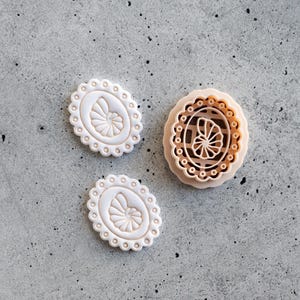May include: Two small, oval-shaped white clay impressions featuring a nautilus shell design with a scalloped border, alongside one larger, round, light tan clay cutter with a grid pattern and a central nautilus shell motif.