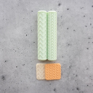 May include: Two cylindrical mint green rollers featuring a vertical herringbone texture, positioned above two small square tiles, one beige and one orange, both embossed with a matching knit-like pattern, arranged to form an exclamation point.