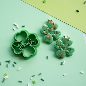 May include: Two green four-leaf clover charms with gold accents and the word "lucky" written on them. A green four-leaf clover cookie cutter is also shown.