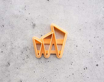 Puede incluir: Un cortador de galletas de plástico color melocotón con un diseño geométrico abstracto que presenta múltiples secciones angulares y puntas afiladas. El objeto tiene un centro hueco y un borde elevado para marcar formas en la masa.