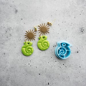 May include: A pair of earrings featuring lime green coiled chameleon charms suspended from gold-toned sunburst studs, accompanied by a single light blue plastic cookie cutter shaped like a coiled chameleon with a detailed spiral design.