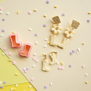 Op de afbeelding: Een paar gele en witte gepatroneerde laarzenoorbellen met een bloemenbedel. Een tweede paar oorbellen heeft een enkele gele en witte gepatroneerde laars. De oorbellen bevinden zich op een lichtgele achtergrond met sprinkels.