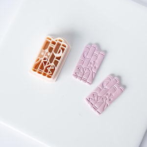 Puede incluir: Un juego de cortadores de galletas marrón claro y rosa. El juego incluye un cortador rectangular con un diseño geométrico y dos galletas rosas con el mismo diseño. El diseño incluye una luna creciente y estrellas.