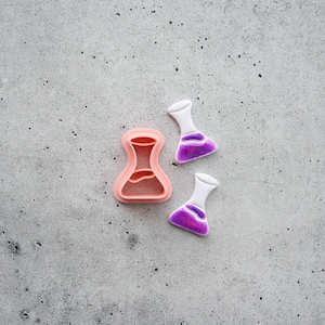 May include: A peach-colored plastic flask-shaped cookie cutter positioned next to two small, white-topped flask-shaped charms or pendants featuring a translucent purple liquid-like center with a visible bubble inside each piece.