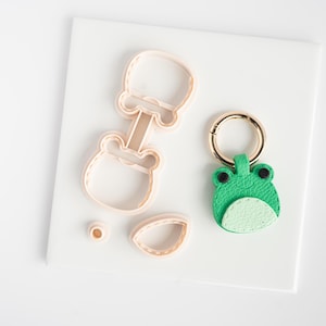 May include: A light pink plastic cookie cutter shaped like a frog, a small circular cutter, a teardrop-shaped cutter, and a finished green and light green faux leather frog keychain with a gold-toned metal ring and a lobster clasp attachment.
