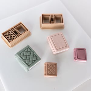 May include: Set of six book-shaped cookie cutters, including a plain design, a design with a diamond pattern, and a design with a grid pattern. The cutters are made of a light brown plastic and are arranged on a white surface.