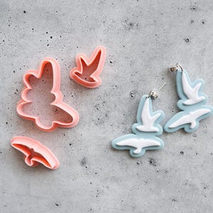 May include: A pair of light blue and white bird-shaped earrings featuring a layered design, alongside three pink plastic cookie cutters in matching bird shapes. The earrings have silver-toned metal posts and backings for attachment.