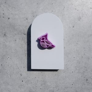 Puede incluir: Un broche con forma de pájaro morado con intrincados patrones de líneas blancas en su superficie, centrado en una tarjeta de presentación blanca y lisa con forma de arco. El diseño del pájaro incluye una cresta puntiaguda y un pico.