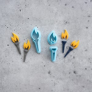 May include: A collection of torch-shaped items including two gray torches with yellow flames, two light blue plastic cookie cutters in the shape of a torch and a flame, and two dark blue torches with yellow flames arranged on a gray surface.