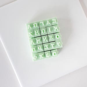 May include: A collection of pale mint green square stamps featuring embossed Greek alphabet letters. The set includes individual blocks arranged in a grid, each displaying a distinct character such as Alpha, Beta, Gamma, Delta, Epsilon, Zeta, Eta, Theta, Iota, Kappa, Lambda, Mu, Nu, Xi, Omicron, Pi, Rho, Sigma, Tau, Upsilon, Phi, Chi, Psi, and Omega.